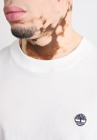 Camiseta de algodón blanca con cuello redondo acanalado, adornada con un pequeño logotipo azul de un árbol en el lado izquierdo cerca del cuello.