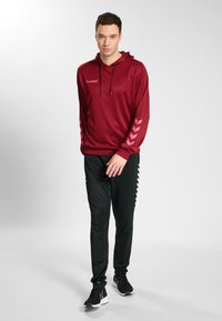 Bordowa bluza z długim rękawem i kapturem, ze sznurkiem i różowymi akcentami w kształcie jodełki; zestawiona z czarnymi sportowymi spodniami z bocznymi paskami.