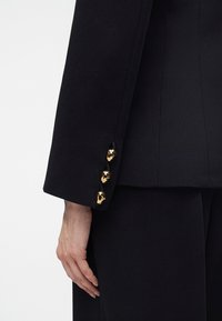 Personne portant une veste de costume noire avec trois boutons dorés brillants sur la manche, main détendue le long du corps devant un fond uni.