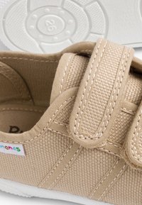 Pantofila din pânză bej cu curea Velcro ajustabilă, talpă din cauciuc alb și cusături texturate. Prezintă un logo colorat pe lateral.