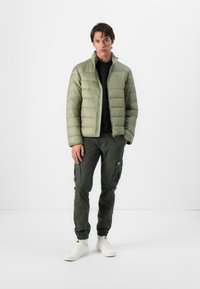 Lichtgroene pufferjack met een ritssluiting aan de voorkant en verticale stiksels, gedragen over een zwart shirt, gecombineerd met donkergroene cargo-broeken en witte sneakers.