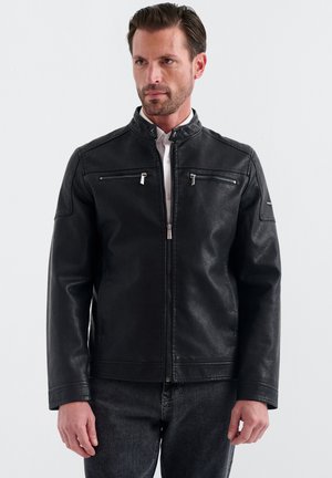 Mann trägt eine schwarze Lederjacke mit Reißverschluss und zwei horizontalen Brustreißverschlüssen, steht vor einem einfarbigen hellen Hintergrund.