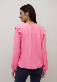 Blouse rose vif avec accents volantés aux épaules, manches longues et dos froncé. Texture lisse, ourlet légèrement courbé, associée à un jean foncé.