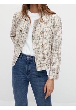 Woman wearing a cream tweed jacket with gold buttons, white top, and blue high-waisted jeans, standing against a plain background.