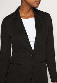 Blazer noir texturé avec une fermeture à un seul bouton, des revers crantés et deux poches avant. Porté sur un haut blanc.