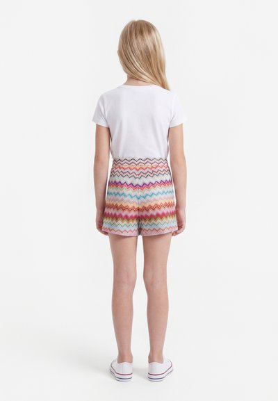 Jeune fille aux longs cheveux blonds portant un t-shirt blanc, un short multicolore à motifs zigzag et des baskets blanches, debout de dos.