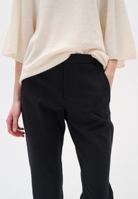 Maglione lavorato a maglia color beige chiaro abbinato a pantaloni neri su misura. I pantaloni hanno una texture liscia e tasche laterali, creando un aspetto elegante.