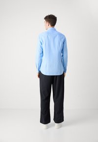 Camicia button-up di un blu chiaro con una texture liscia, abbinata a pantaloni neri a righe sottili. Il modello è in piedi di spalle, indossando scarpe bianche.