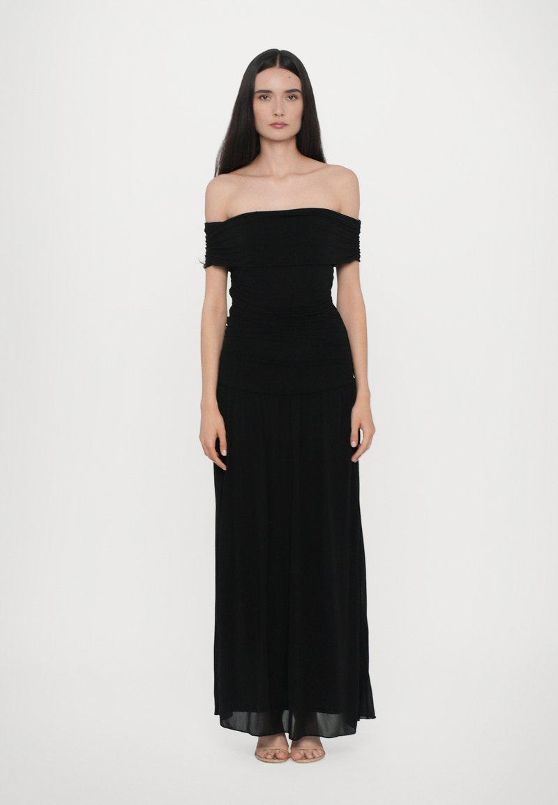 Zwarte off-shoulder jurk met geplooide textuur bij de buste, vloeiende maxi lengte en een gladde afwerking. Model staat tegen een witte achtergrond.
