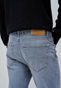 Jean en denim bleu clair avec une coupe droite, arborant une étiquette en cuir beige au dos à la taille. Les poches arrière présentent un design cousu.