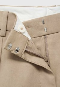 Gros plan de la taille d’un pantalon beige avec fermeture éclair partiellement ouverte, bouton blanc et deux crochets métalliques sur la ceinture en tissu.