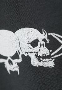 Crni materijal s bijelom grafikom koja prikazuje dva stilizirana lobanja, detaljno izrađena s očnjacima, šupljinama za oči i teksturiranim sjenčanjem.