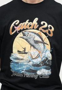 Μεγάλο ψάρι που πηδάει έξω από το νερό κοντά σε βάρκα με ψαρά, κείμενο "Catch 23" και "Jordan Fishing Club" σε μαύρο πουκάμισο.