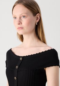 Robe noire côtelée avec décolleté dégageant les épaules, boutons décoratifs à l'avant et bords festonnés. Fabriquée dans un tissu texturé.