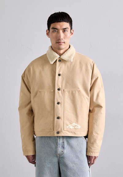 Veste en toile kaki avec de grandes poches avant, col en fausse fourrure beige, fermeture à boutons et détail de logo blanc à l'ourlet. Coupe décontractée et confortable.