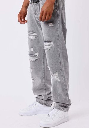Personne portant un jean gris ample déchiré et des baskets blanches Nike, debout devant un fond uni clair.