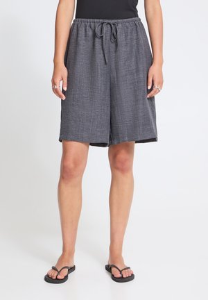 Person iført løse grå shorts med snøre og sorte flipflops står på en ensfarvet hvid baggrund.