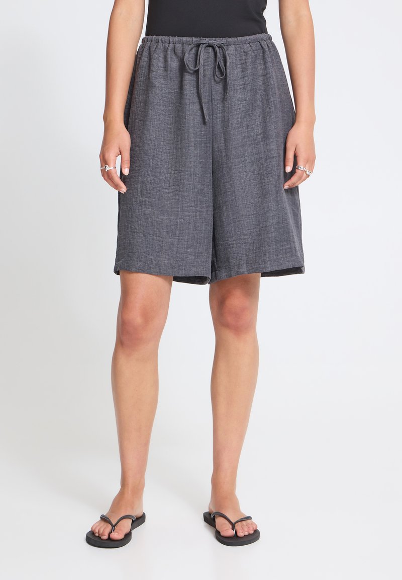 Personne portant un short gris ample avec cordon de serrage et des tongs noires, debout sur un fond blanc uni.