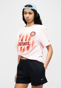 Γυναίκα που φοράει ανοιχτό μπλε καπέλο, ροζ παλ μπλουζάκι Nike με την επιγραφή "Just Do It" και μαύρα σορτς Nike, με τα χέρια στις τσέπ ες, στέκεται μπροστά σε απλό φόντο.