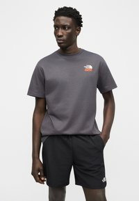 Νεαρός άνδρας που φοράει γκρι μπλουζάκι The North Face και μαύρο σορτς, στέκεται με το ένα χέρι στην τσέπη, κοιτώντας προς τα πλάγια.