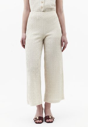 Vrouw die crèmegekleurde, hoogtaillotte, wijd-uitlopende broeken met structuur draagt, gecombineerd met donkerbruine open schoenen met gouden gespen op een witte achtergrond.