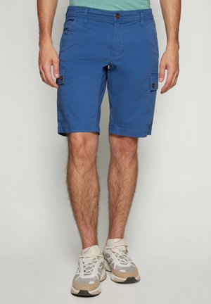 Mand iført blå cargoshorts og hvide sneakers står mod en ensfarvet baggrund.