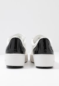 Baskets blanches avec une tige texturée et des accents noirs brillants au talon. Semelle compensée, coutures blanches et détails du logo visibles.