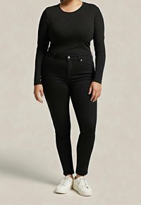 Personne portant un haut noir ajusté à manches longues et un jean skinny noir associé à des baskets blanches, debout devant un fond neutre.