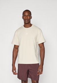 T-shirt beige à manches courtes avec un col rond et un logo trois rayures subtil en blanc sur la poitrine, associé à un short bordeaux.