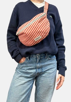 Personne portant un jean bleu clair et un pull bleu marine, avec un sac bandoulière rayé rouge et blanc avec fermeture éclair et étiquette logo.