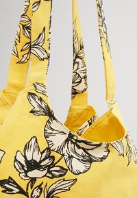 Borsa in tela gialla con stampa floreale nera, dotata di due spallacci e una texture morbida. L'interno è completamente giallo senza scomparti.