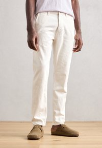 Vita bomullbyxor med rak passform, utrustade med bälteshällor, sidofickor och en texturerad yta. Parades med bruna mockaskor.