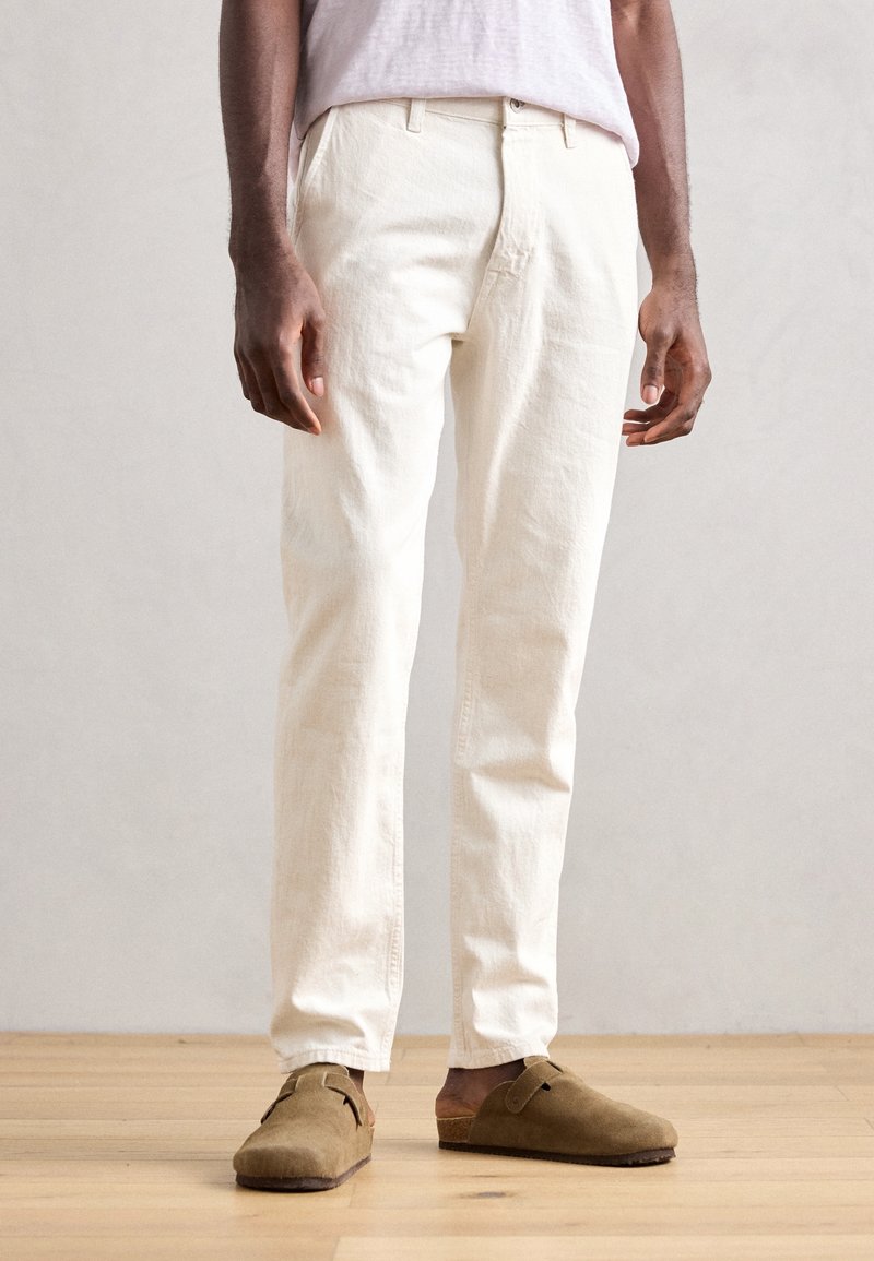 Vita bomullbyxor med rak passform, utrustade med bälteshällor, sidofickor och en texturerad yta. Parades med bruna mockaskor.