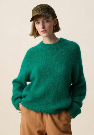 Femme portant un pull vert en matière pelucheuse, un pantalon beige, une casquette à imprimé léopard et des boucles d'oreilles dorées en forme d'anneau, debout avec les mains dans les poches.