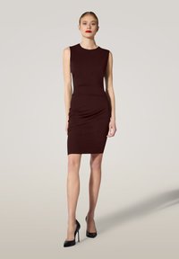 Frau in einem ärmellosen, dunkelbraunen, figurnahen knielangen Kleid und schwarzen High Heels, die vor einem schlichten hellen Hintergrund steht.