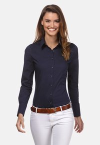 Camicia aderente blu navy con maniche lunghe, colletto button-down, bottoni frontali e polsini a contrasto, abbinata a pantaloni bianchi e una cintura marrone.