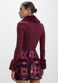 Maglione a coste bordeaux con colletto e polsini in pelliccia sintetica abbinato a una mini gonna a fantasia con forme audaci in rosso, rosa e viola.