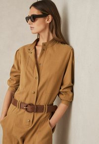 Brun bomulls jumpsuit med hög krage, tryckknappar och två framfickor, åtsmitande i midjan med ett flätat brunt läderbälte.