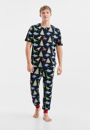 Ensemble de pyjama pour homme comprenant un tissu noir avec des motifs de dinosaures colorés et de sapins de Noël. Haut à manches courtes et pantalon long avec des poignets rouges.