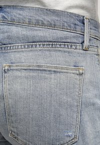 Pantalones vaqueros de mezclilla azul claro con una textura ligeramente desteñida, costuras visibles y un solo bolsillo trasero. Presentan una cinturilla estándar y trabillas para cinturón.