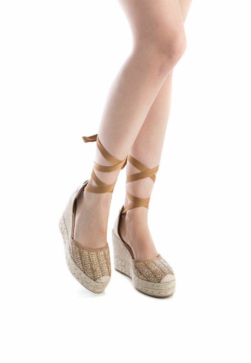 Alpargatas de cuña con parte superior de yute trenzado, ataduras de tela marrón claro en el tobillo y suela texturizada. Los colores incluyen beige y marrón claro.