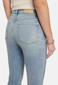 Jeans en denim bleu clair avec une taille haute, cinq poches et une étiquette en cuir sur le dos de la taille. Texture lisse, design ajusté.