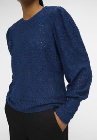 Blusa blu navy con motivo in pizzo estruturato e intricato, e maniche lunghe. Presenta un colletto a giro e un dettaglio di spalla leggermente gonfiata.