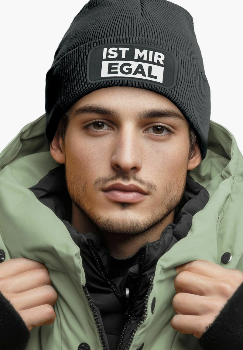 Black knitted beanie with "IST MIR EGAL" label, paired with a green puffer jacket. Close-up view of model's face and hands.