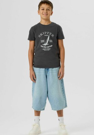 Usmívající se chlapec stojící, oblečený v tmavě šedém tričku s grafikou plachetnice a světle modrých dlouhých džínových kraťasech na jednoduchém světle pozadí.