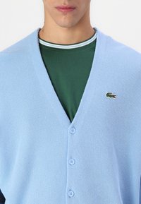 Cardigan lavorato a maglia di colore blu chiaro con scollo a V, caratterizzato da chiusure a bottoni e un piccolo logo di coccodrillo ricamato in verde, indossato sopra una camicia verde scuro.