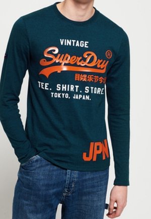 Chemise à manches longues vert sarcelle foncé avec texte orange de style vintage, incluant "SuperDry" et "JPN", avec un col rond et des poignets côtelés.
