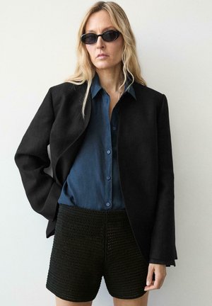 Lyshåret kvinde iført sorte solbriller, sort åben jakke, blå skjorte med knapper og teksturerede sorte shorts poserer mod hvid baggrund.