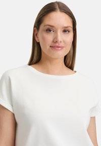 Biała bluzka z krótkim rękawem, okrągłym dekoltem, prążkowaną fakturą i podwiniętymi mankietami. Gładki materiał z luźnym krojem.