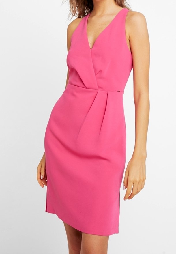 Femme portant une robe rose sans manches, ajustée, longueur genoux, avec un col en V et des plis à la taille, sur un fond blanc uni.